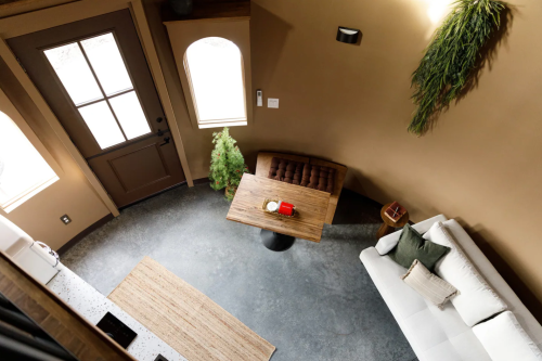 A cozy interior view featuring a wooden table, sofa, and a small tree, with natural light from a door and windows.